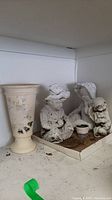 Three white garden figures with a small container, and cream-colored tall vase with floral design on a shelf.