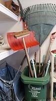 Garden tools in green Ottawa City Services waste bin including large leaf rake, orange shovel, broom and more
