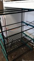 Frame of greenhouse showing four metal wire shelves and green metal bars for structure