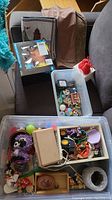 Multiple plastic bins filled with assorted small toys, dolls, and miniatures arranged on a couch along with some place mats and fabric items.