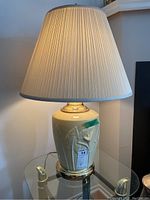 Full view of large yellow ceramic lamp on glass table, with pleated cream lampshade and brass base trim