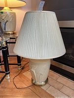 Full view of large yellow ceramic lamp with brass trim base and pleated cream lampshade sitting on floor near fireplace.
