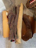 Photo showing rolled textiles including a woven tapestry, a yellow yoga mat, a fur textile, and a rolled leather hide on carpet.