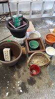 Various plant pots in assorted sizes, plastic, terra cotta, stone, and woven basket shown on floor.