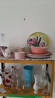 Photo of shelf showing two plates, pink trivet, Royal Oak teapot, and various glass and ceramic vases including clear, blue, white, and red patterns.