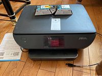 HP Envy 5660 printer showing top and front with power button and paper output tray, along with several HP 62 ink cartridge boxes on top, placed on hardwood flooring.