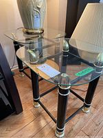 Two glass top side tables with black cylindrical legs and gold-tone metal trim, photographed on wooden floor beside a lamp and other furniture.