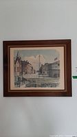 Full view of framed print showing street scene and buildings with power lines and a river dock area, framed in a brown wood frame.