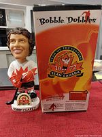 Bobby Clarke bobblehead figure standing on white base with nameplate, wearing white and red Team Canada jersey and black hockey gear, holding hockey stick. Original box beside figure features Team of the Century 1972 Team Canada logo and Bobble Dobbles branding.