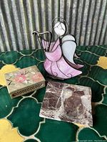 Three decorative items on green patterned surface: stained glass angel, stone box with floral inlays, and marble piece.