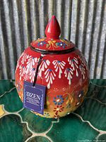 Front view of red round ceramic vase with floral hand-painted decoration, showing attached brand tag