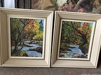 Pair of framed landscape paintings on board in cream frames, showing autumn scenes with streams and trees.