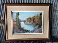 Full view of the vintage framed landscape painting showing trees by a water body with an autumn palette, in a dark brown wooden frame with fabric-textured matting.