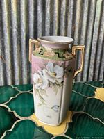 Front view of hexagonal vintage Nippon vase showcasing hand painted white flowers on pink and cream background with geometric green and gold border near the rim and gold accented handles.