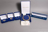 Five Camrose & Kross blue velvet presentation boxes containing two brooches (silver filigree, gold swirl), a pair of earrings, a necklace, and a bracelet, plus certificate of authenticity displayed in front.