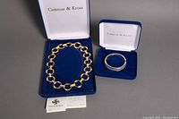 Full view of Camrose & Kross gold-tone necklace and hinged bangle bracelet in original blue velvet-lined boxes, accompanied by certificate cards.