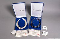 Two original blue velvet-lined Camrose & Kross boxes containing costume jewelry sets: one with faux pearl necklace and one with black crystal necklace and earrings, accompanied by certificates of authenticity and small branded boxes.
