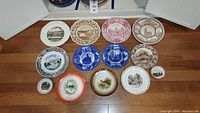 Overview of collection of 14 antique collector plates arranged on the floor, showing various designs including historical buildings, advertising, and animal illustrations