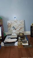 Overview photo showing the large bird canvas artwork leaning against the wall on a table holding various picture frames and decorative plaques piled and arranged on the floor beneath it.