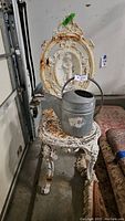 Side view of the white cast iron chair with ornate cherub relief and galvanized watering can on seat, showing rust and peeling paint.