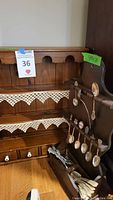 Wooden spoon display rack with lace trimmed shelves and small drawers, holding spoons and flatware.