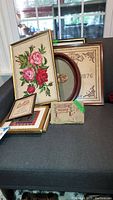 Group of eight framed needlepoints displayed on dark surface and dog door stop in front showing variety of sizes and frame types.