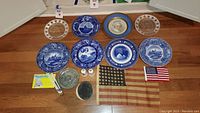 Full view of the lot including various blue and white decorative George Washington plates, portrait plates, two salt and pepper shakers, a metal medallion, and three historical American flags (48-star and Bicentennial).