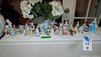 Wide shot showing 21 vintage porcelain figures arranged on a ledge. Many small detailed figures showing children and animals, including a prominent Cinderella figure in a blue dress on the right.