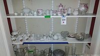 Two shelves displaying a collection of miniature items including small teapots, creamers, sugar bowls, cups, saucers, and Wedgwood plates. Items vary in design and decorations with some having floral patterns and gold trim. Some cup handles appear missing and some pieces show wear.