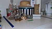 Wide view of the lot showing multiple perfume bottles, striped hat box with straw hat, and the fabric pin cushion on a white surface