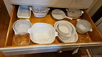 Top view of drawer displaying white milk glass and Corning Ware dishes, bowls, and clear glass lids including Pyrex measuring bowls and a casserole dish with cornflower pattern.