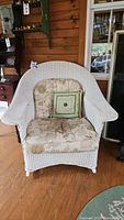 Front view of white reed wicker rocking chair with floral cushions and a green decorative pillow on wooden floor indoors.