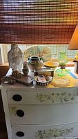 Overall view of the lot on white floral dresser includes Buddha statue, deer plaque, hobnail oil lamp, vase, rock, marble, plate, and bowl under window light.