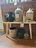 Twelve vintage pottery pieces arranged on and under a small white table, showing variety of shapes and sizes including jugs and crocks with tags indicating lot numbers.
