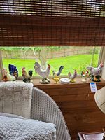 Wide view of 11 vintage porcelain and ceramic rooster figurines in various sizes and colors displayed on window ledge with an outdoor garden background