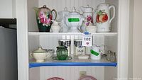 Upper shelf with assorted pitchers and lidded bowls including floral and fruit motifs, labeled with lot tag.