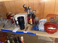Overview of barware lot on wooden shelf showing stemware, clear ice bucket, ceramic pitcher, black plastic cups, and bar tools.