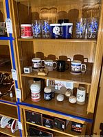 Shelf display showing multiple coffee mugs and glasses including Budweiser mug, Orioles and Colts themed glass tumblers, and signed baseballs on lower shelf.