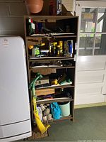Front view of six-tier wooden bookshelf containing various tools and household items on shelves, refrigerator to left and door to right.