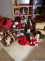 Photo showing various Christmas stockings, figurines including Santa Claus and Precious Moments pieces, snowman decoration, poinsettia flowers, and basket on floor and shelf.