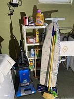 Photo shows basement area with blue Hoover WindTunnel upright vacuum cleaner with bag, two ironing boards with floral covers, mop, broom, dustpan, and shelves with cleaning chemicals.