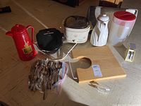 Overview photo of all items including red coffee carafe, crock pot, electric pot, pitcher, cutting board, utensils, and grater on table.