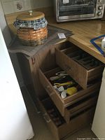 View of kitchen corner showcasing open drawers containing kitchen utensils, flatware organizer with Cambridge stainless flatware, nut crackers, and tools, with a woven cookie jar on the counter.