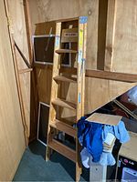 Wooden step ladder standing in a basement corner with laundry and other stored items nearby. Ladder has safety labels visible.