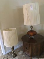 Two vintage table lamps placed in a corner. One lamp with glass brown-toned base and fabric shade on a wooden hexagonal table, the other with brass-toned base and fabric shade placed on the floor.