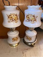 Two matching vintage glass globe lamps standing side by side. Each lamp shows detailed yellow and brown floral patterns on both the upper globe with ruffled edge and the lower base glass portion. Brass colored metal accents are visible on the base. Electrical cords attached.