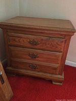 Overall view of three-drawer wooden nightstand with oak finish in corner against wall.