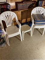 Four white plastic stackable outdoor chairs, one small white plastic side table, and two folded camping chairs in background stacked in a basement setting.