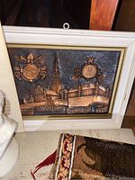 Metal embossed plaque with pope coins and 1983 cityscape in white frame placed on a shelf.