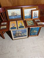 Full view showing 11 framed ship paintings on bench and floor with various sizes and frame styles